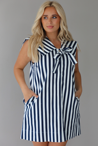 Woman wearing a navy and white striped dress with a bow detail at the neckline indoors against a white wall. The photo shows a closeup of the dress, with the models hand in the pockets on either side. 