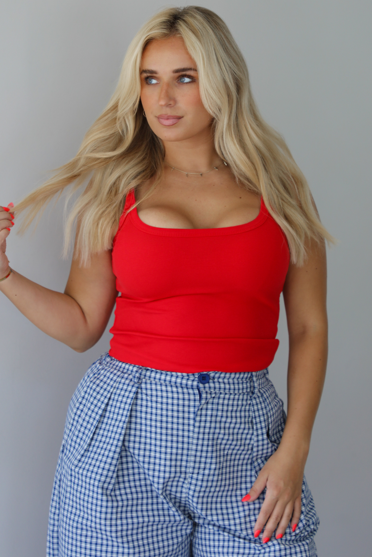 Woman wearing a red tank top high waisted navy and white plaid pants indoors against a white wall. The photo shows a closeup of the tank, which has a ribbed texture. 
