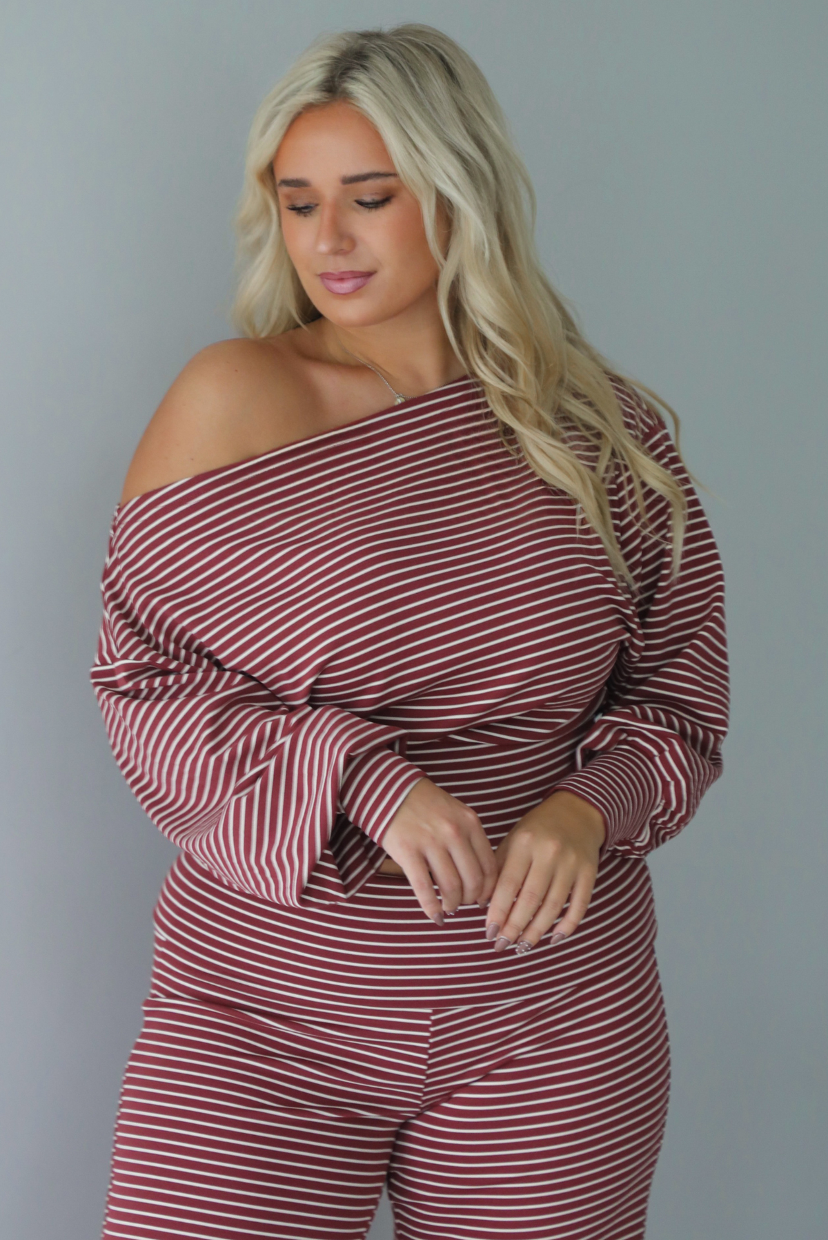 A woman wearing a maroon and white striped two piece matching set featuring a slouchy off the shoulder long sleeved top and mid-rise, folded over waistline wide leg pants, standing indoors against a white wall.