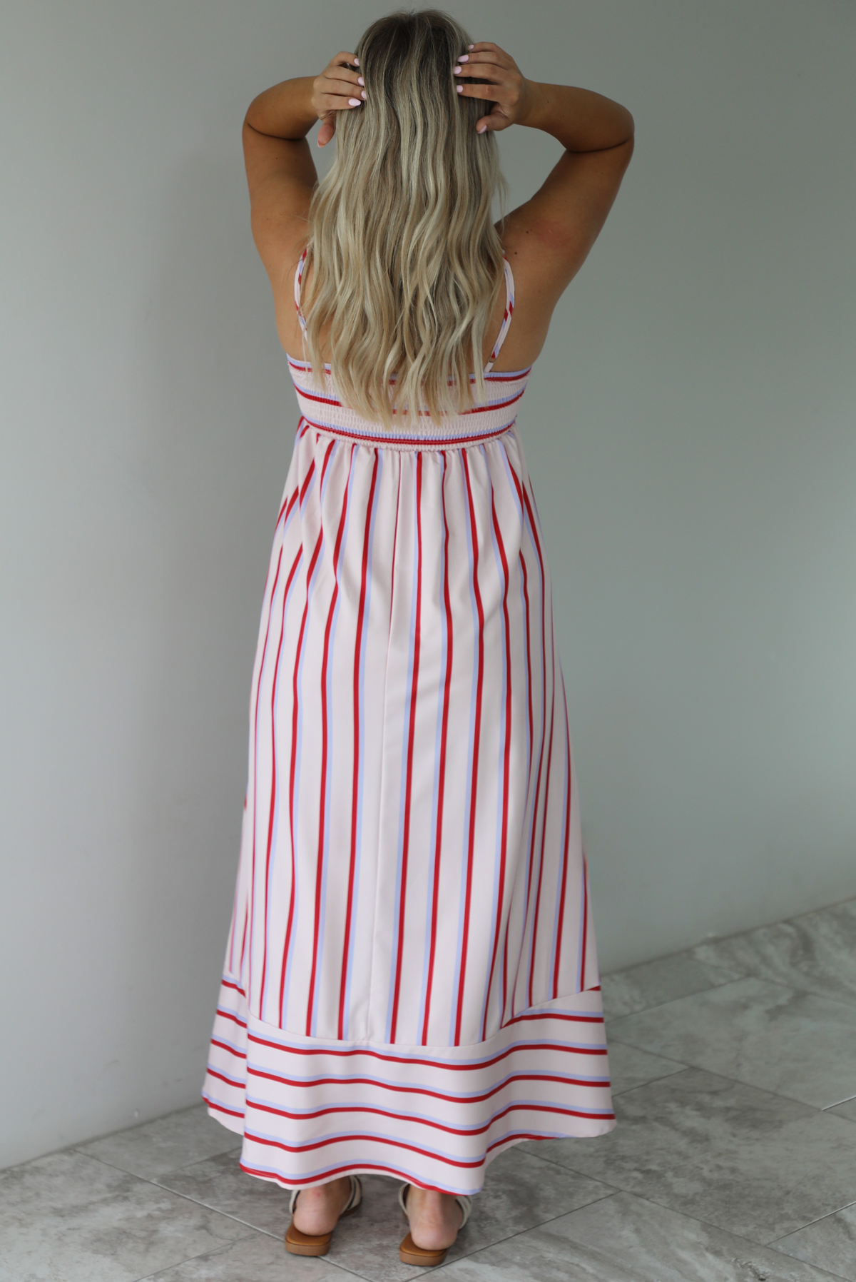 Woman wearing a spaghetti strap, maxi length dress that has a light pink color throughout with a red and light blue striped pattern. The photo is taken indoors against a white wall with white marble flooring. The photo shows the back of the dress, which has adjustable straps and a smocked back. 