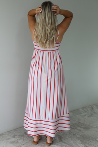 Woman wearing a spaghetti strap, maxi length dress that has a light pink color throughout with a red and light blue striped pattern. The photo is taken indoors against a white wall with white marble flooring. The photo shows the back of the dress, which has adjustable straps and a smocked back. 