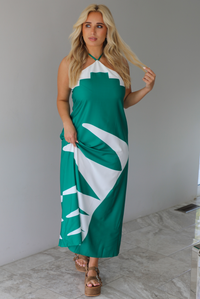 Woman wearing a green and white maxi dress with a halter neckline indoors against a white wall with white marble flooring. 