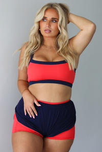 A woman modeling a two-piece athletic set with a bright coral-red and navy blue color-blocked design, standing indoors against a white wall.