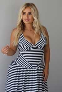 A closeup of a woman modeling a sleeveless, V-neck maxi dress with a black and white horizontal stripe pattern, focusing on a closeup of the bodice, which is a drop waist design, standing indoors against a white wall.