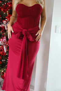 Close up of a woman model wearing a strapless, red holiday maxi length dress. The dress has a corset style bodice, a tie detail at the waistline, and a stretchy, satin textured maxi length skirt. The photo is taken inside a modern home in front of a decorated Christmas tree. 