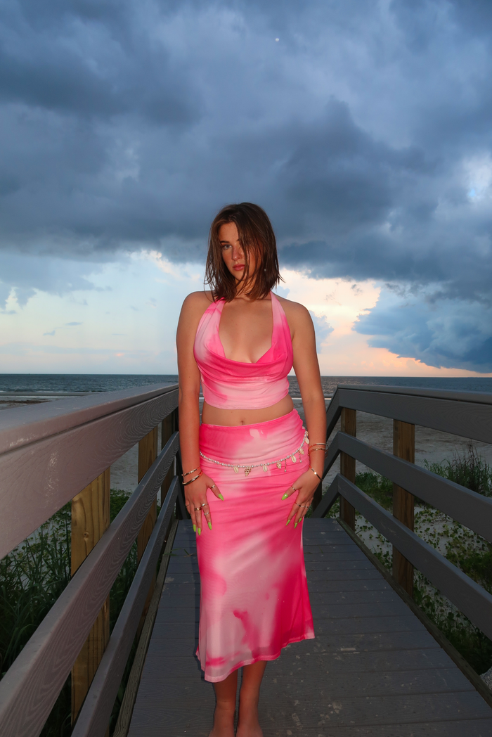 A woman stands on a wooden boardwalk at the beach wearing a bright pink and light pink tie-dye two-piece set consisting of a halter crop top and a midi skirt. The background features a dramatic cloudy sky and the ocean.
