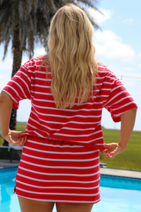 Woman in a red, light blue, and white striped two piece set featuring an oversized top and mini skirt outdoors in front of a pool with palm trees in the background. The photo shows the back of the set.