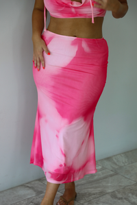 A woman wearing a two-piece set featuring a bright pink and light pink tie-dye pattern, consisting of a halter crop top and a matching midi skirt. 