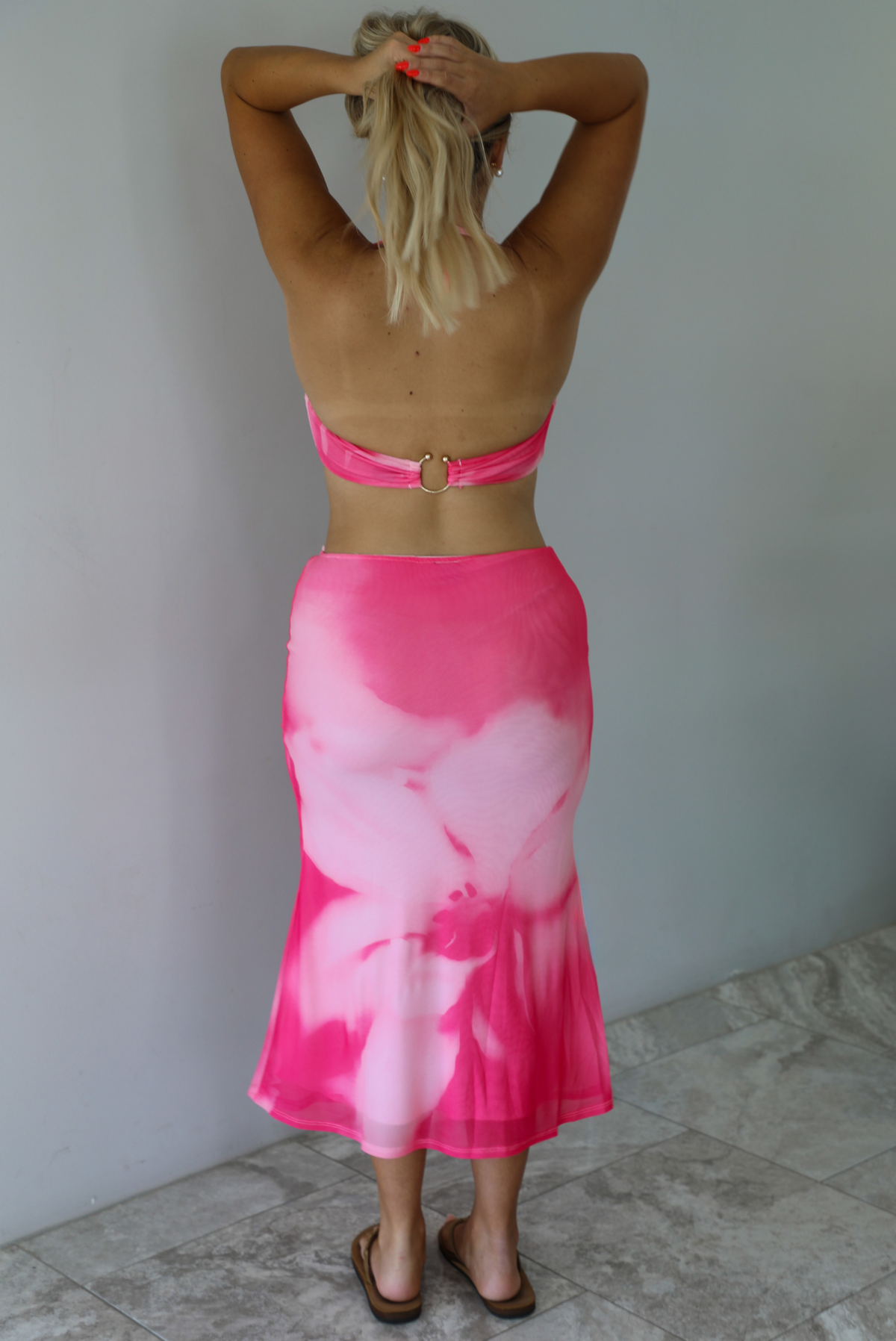 A woman viewed from behind wearing a bright pink and light pink tie-dye halter top and matching high-waisted skirt, standing against a white wall.