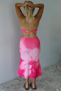 A woman viewed from behind wearing a bright pink and light pink tie-dye halter top and matching high-waisted skirt, standing against a white wall.