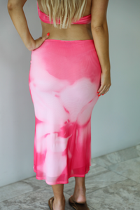 A woman shown from the back wearing a bright pink and white tie-dye skirt with a matching halter top, standing indoors against a white wall with white marble flooring.