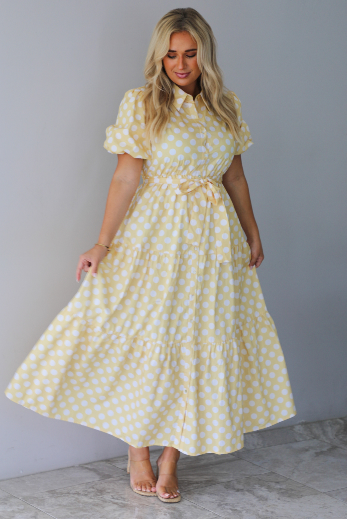 A woman wearing a yellow and white polka dot midi dress with puff sleeves and a tie waist and functional button closures along the front, standing indoors against a white wall with white marble flooring. 