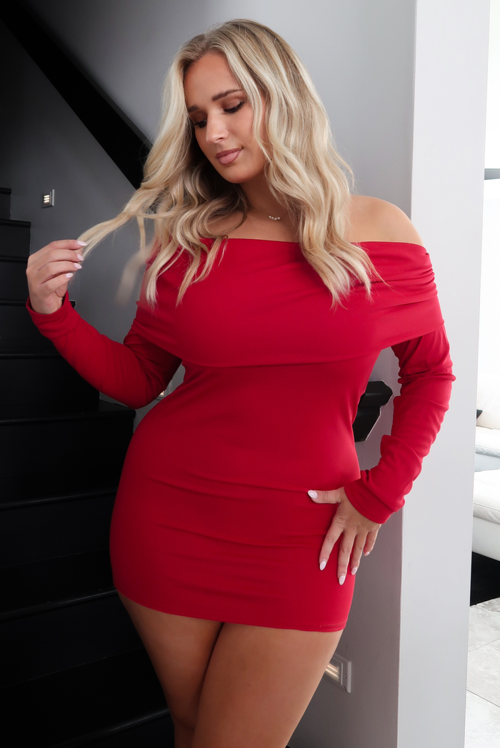Woman model wearing a red mini dress in a modern white and black home interior. The dress has an off the shoulder design that folds over at the upper half of the arms, has long sleeves, and a mini length design. The photo shows a close up of the dress with the folded over off the shoulder neckline, long sleeves, and fitted fit. The photo is taken in front of a black staircase, with a glimpse of a white couch in the background. 