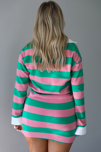 Woman wearing a two piece pink and green striped set featuring a collared top and fitted skirt. The photo is taken indoors in front of a white wall and shows the back of the set. 