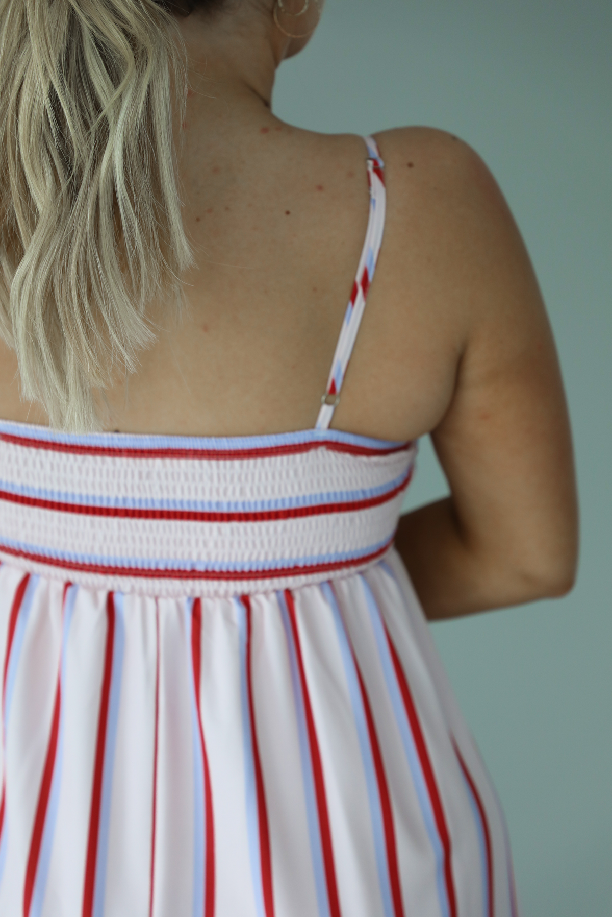 Woman wearing a spaghetti strap, maxi length dress that has a light pink color throughout with a red and light blue striped pattern. The photo is taken indoors against a white wall and shows the back of the dress, which has adjustable straps and a smocked back. 