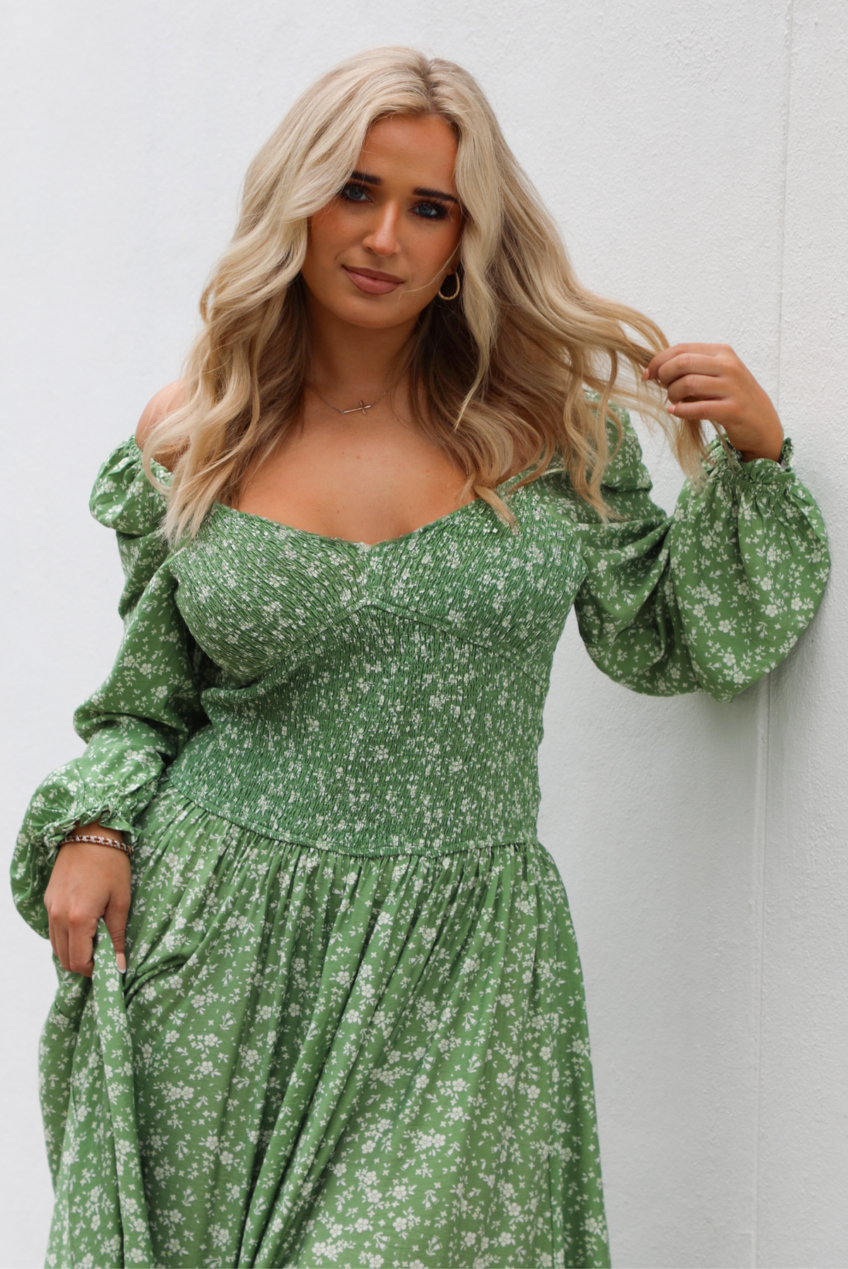 Woman model wearing a green maxi length dress that has a white floral print pattern, long sleeves, smocked bodice, and a flowy skirt. The model is holding the skirt up on one side. The photo is taken outdoors against a white wall.  The photo shows a closeup of the bodice and long sleeves. 