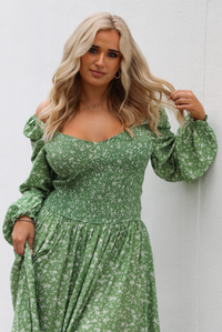Woman model wearing a green maxi length dress that has a white floral print pattern, long sleeves, smocked bodice, and a flowy skirt. The model is holding the skirt up on one side. The photo is taken outdoors against a white wall.  The photo shows a closeup of the bodice and long sleeves. 