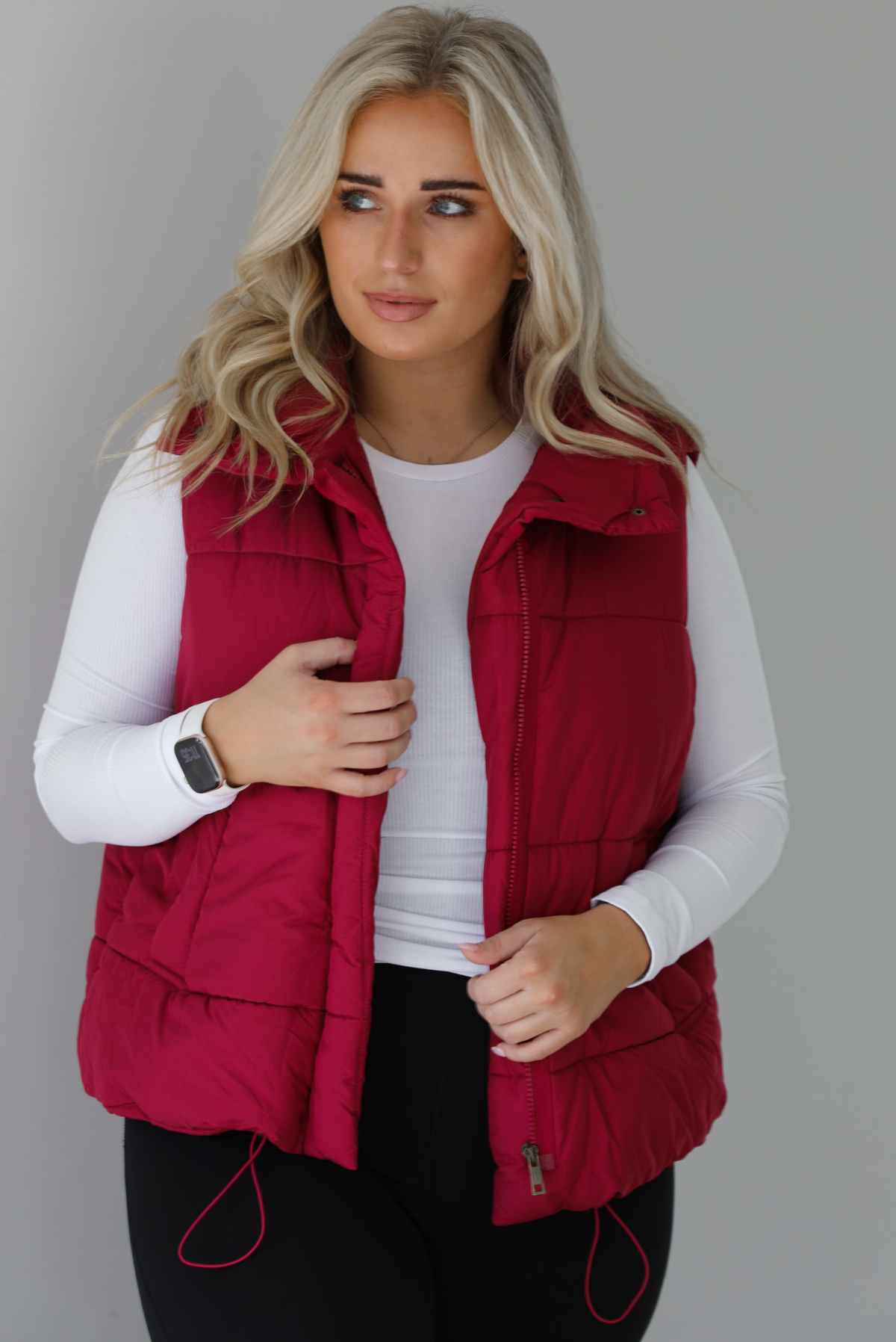 Woman wearing a wine red puffer vest, white long-sleeve shirt, and black leggings indoors against a white wall. The puffer vest has a collared design, functional button closures at the neckline, an unzipped closure in the front, and functional pockets on either side.