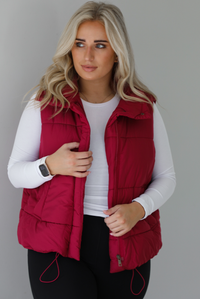 Woman wearing a wine red puffer vest, white long-sleeve shirt, and black leggings indoors against a white wall. The puffer vest has a collared design, functional button closures at the neckline, an unzipped closure in the front, and functional pockets on either side.