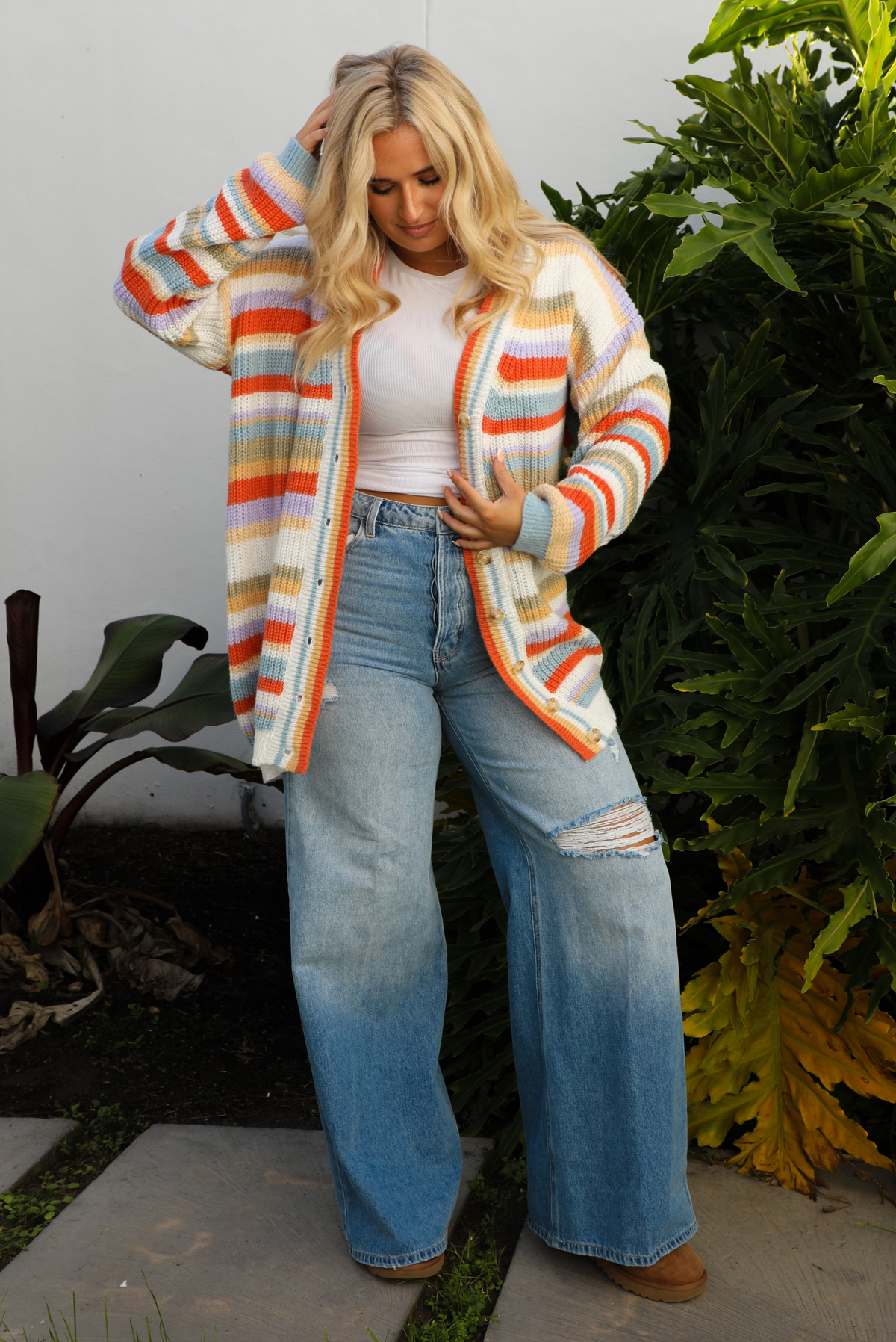Woman model wearing an orange, lavender, yellow, muted green, blue, and white striped oversized cardigan with functional button closures in the front and an oversized fit with a light wash flared hem denim. The photo is taken outdoors against a white wall and greenery in the back. The photo shows the cardigan unbuttoned with a white cropped top underneath.