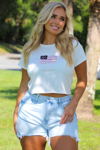 Woman wearing a white baby tee with an embroidered American Flag on the front with light wash denim shorts outdoors with greenery and palm trees in the background. 