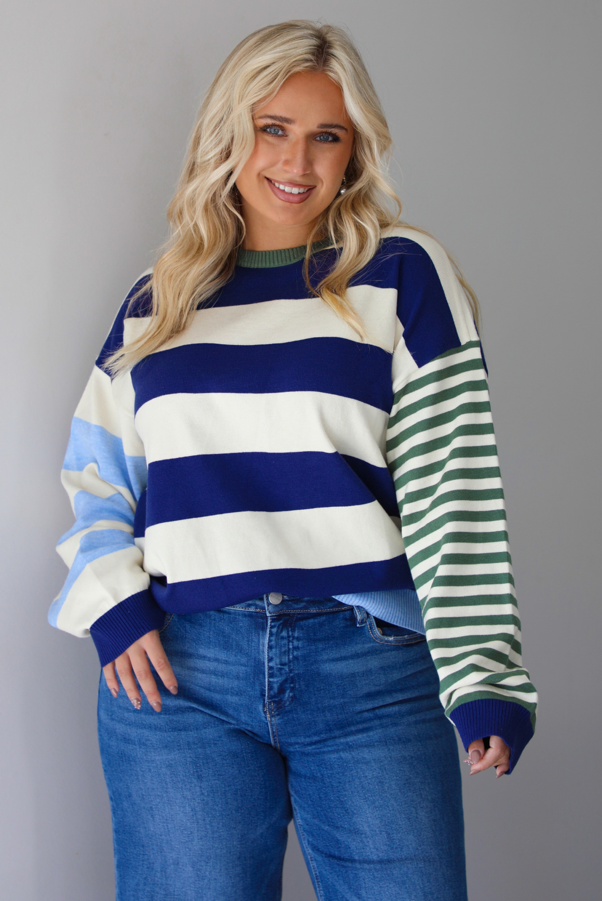 Woman wearing a navy, cream, blue, and green alternating striped design throughout, paired with dark denim jeans, standing indoors against a white wall.