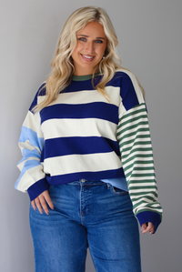 Woman wearing a navy, cream, blue, and green alternating striped design throughout, paired with dark denim jeans, standing indoors against a white wall.
