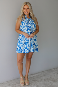 Woman wearing a white and blue print dress with a tree branch and bird pattern throughout indoors against a white wall with white marble flooring. 