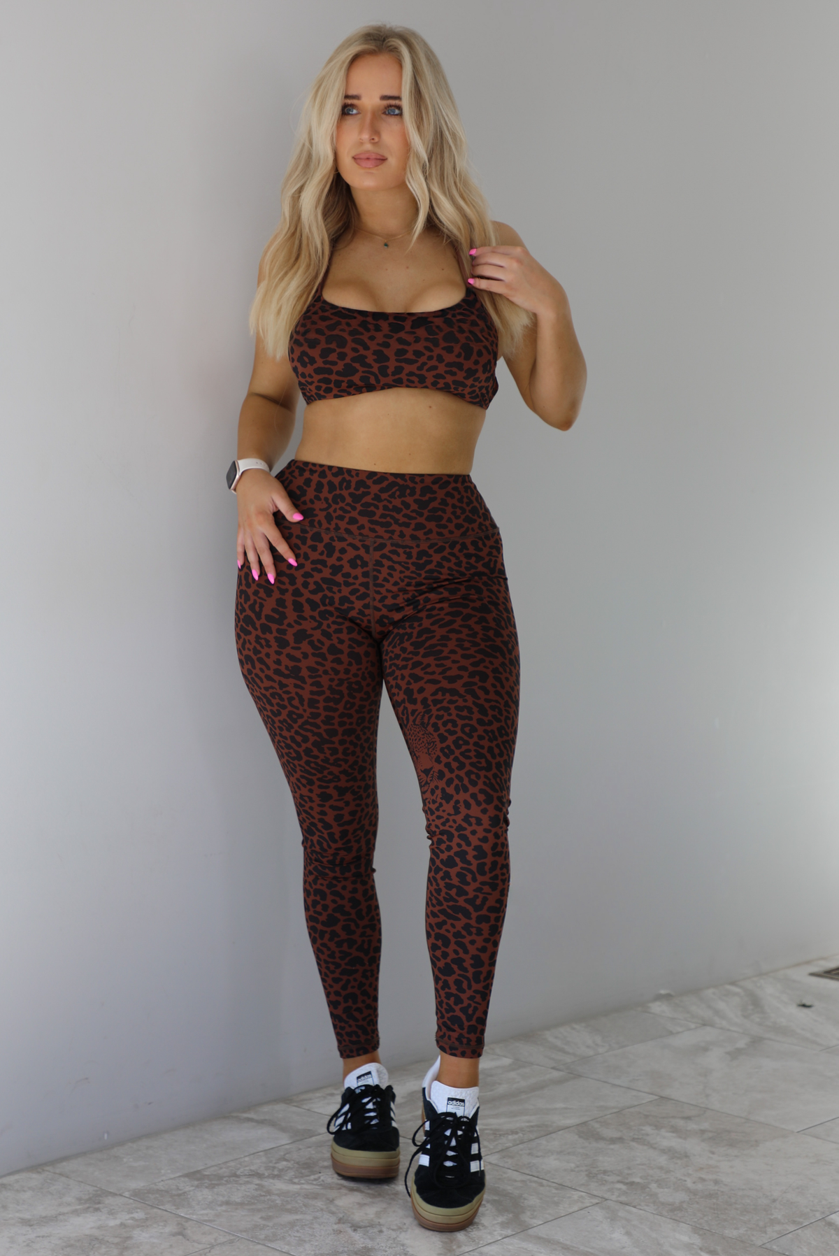 Woman wearing a brown and white leopard print two piece athleisure set that consists of a sports bra and high waisted leggings. The photo is taken indoors against a white wall with white marble flooring. 
