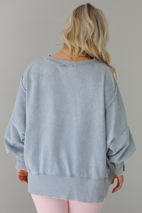 Woman wearing a grey colored oversized sweatshirt with exposed seam details and a high-low design with pink leggings. The photo shows the back of the sweater. The photo is taken indoors against a white wall.