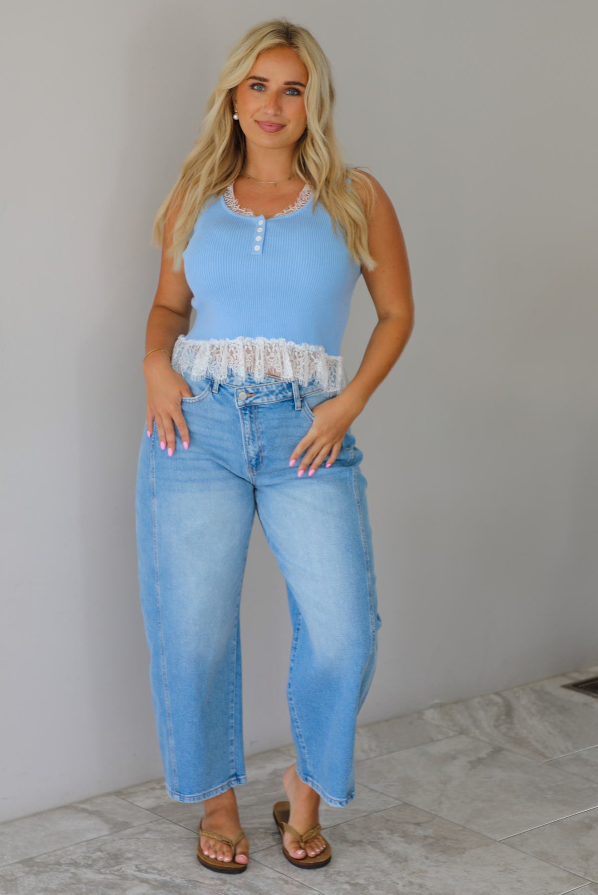 A woman wearing a light blue sleeveless top with a white lace hem and light blue high-waisted cropped jeans, standing indoors against a white wall with white marble flooring. 