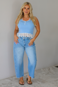 A woman wearing a light blue sleeveless top with a white lace hem and light blue high-waisted cropped jeans, standing indoors against a white wall with white marble flooring. 