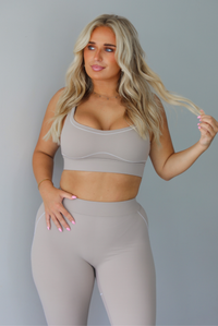 A woman modeling a matching tan activewear set consisting of a sports bra and high-waisted leggings with white stitching details, standing indoors against a white wall.