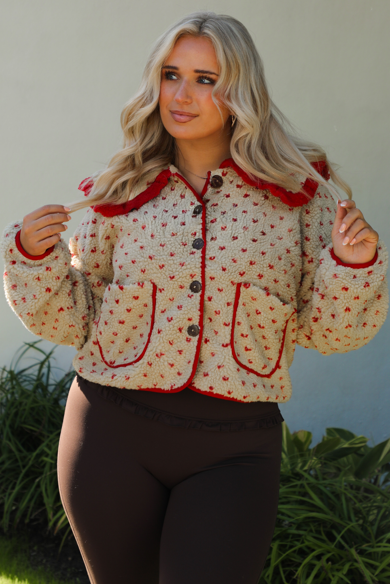Woman model wearing a tan and red sherpa cardigan with a collared neckline and red trim with dark brown ahtleisure pants. The photo is taken outdoors with a white wall and greenery in the back. 