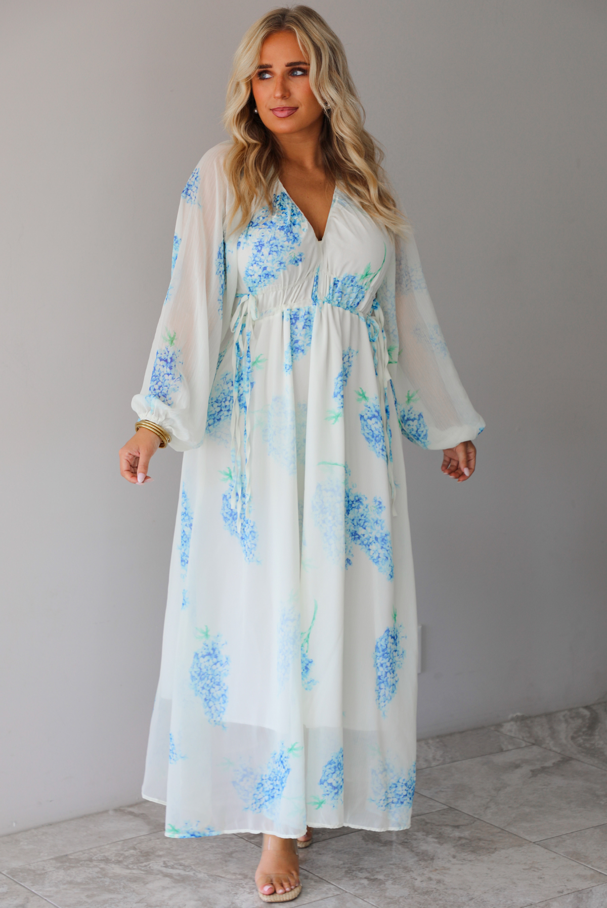 A woman modeling a white maxi dress with a blue floral print and a sheer, flowing overlay, standing indoors against a white wall with white marble flooring. 