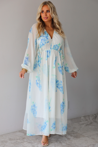 A woman modeling a white maxi dress with a blue floral print and a sheer, flowing overlay, standing indoors against a white wall with white marble flooring. 