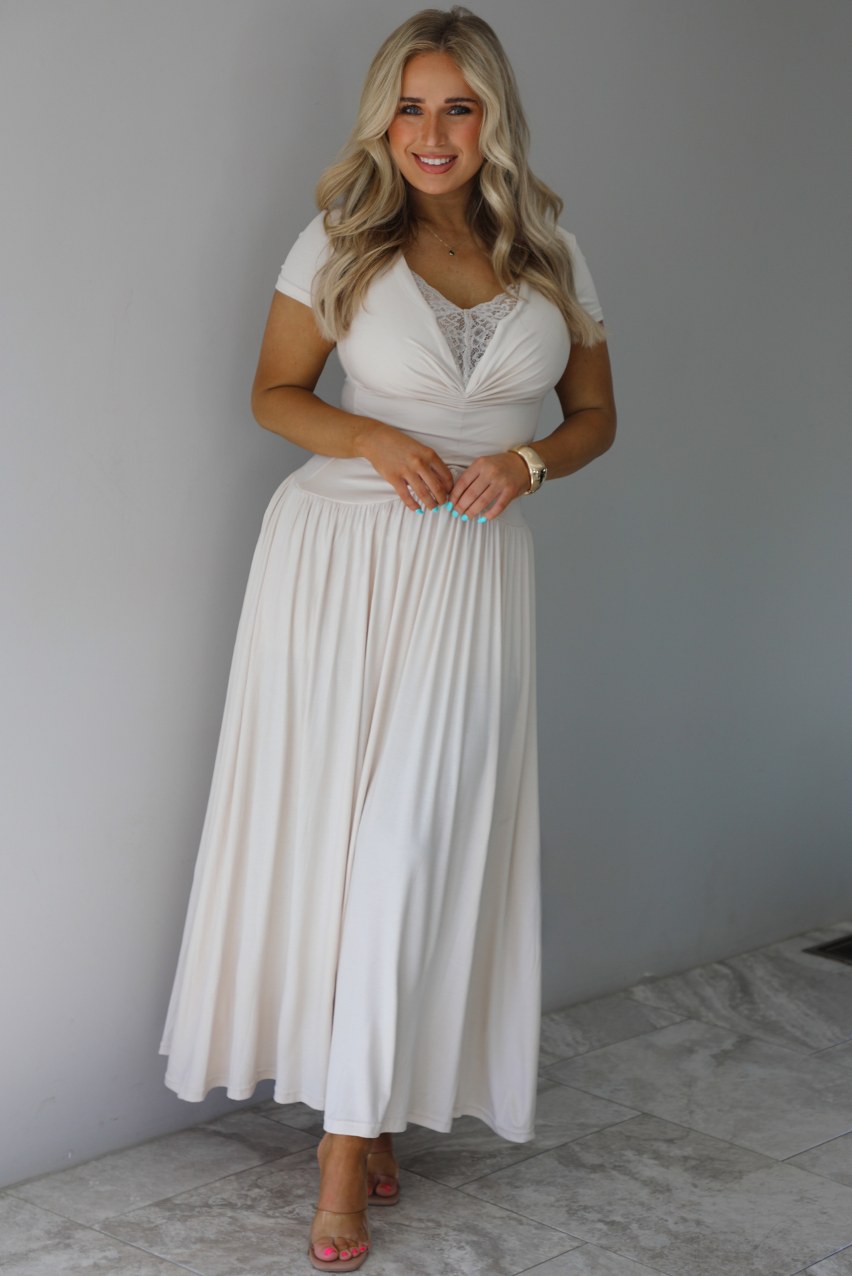 Woman wearing a light sand colored maxi length dress that has a fitted bodice with lace detailing in the center of the bust, a drop waist design, short sleeves, and a flowy skirt. The photo is taken indoors against a white wall with white marble flooring. 