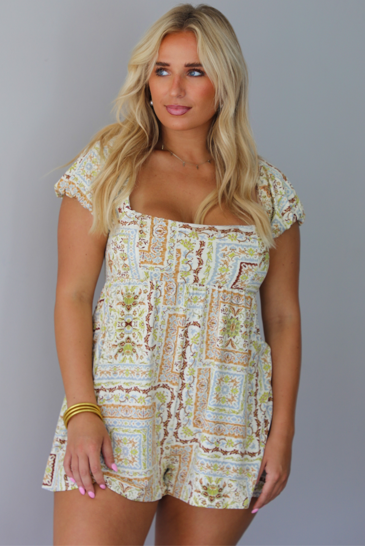 A woman wearing a cream-colored short-sleeve romper with a beige and brown geometric pattern, standing indoors against a white wall.