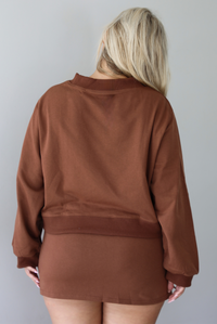 A woman from behind wearing a brown matching two piece set featuring a cropped sweatshirt and mini skirt of the same material, standing indoors against a white wall. 