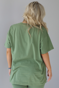 Woman wearing a green two piece set featuring an oversized t-shirt and fitted leggings. The photo shows a closeup of the back of the oversized top. The photo is taken indoors against a white wall.