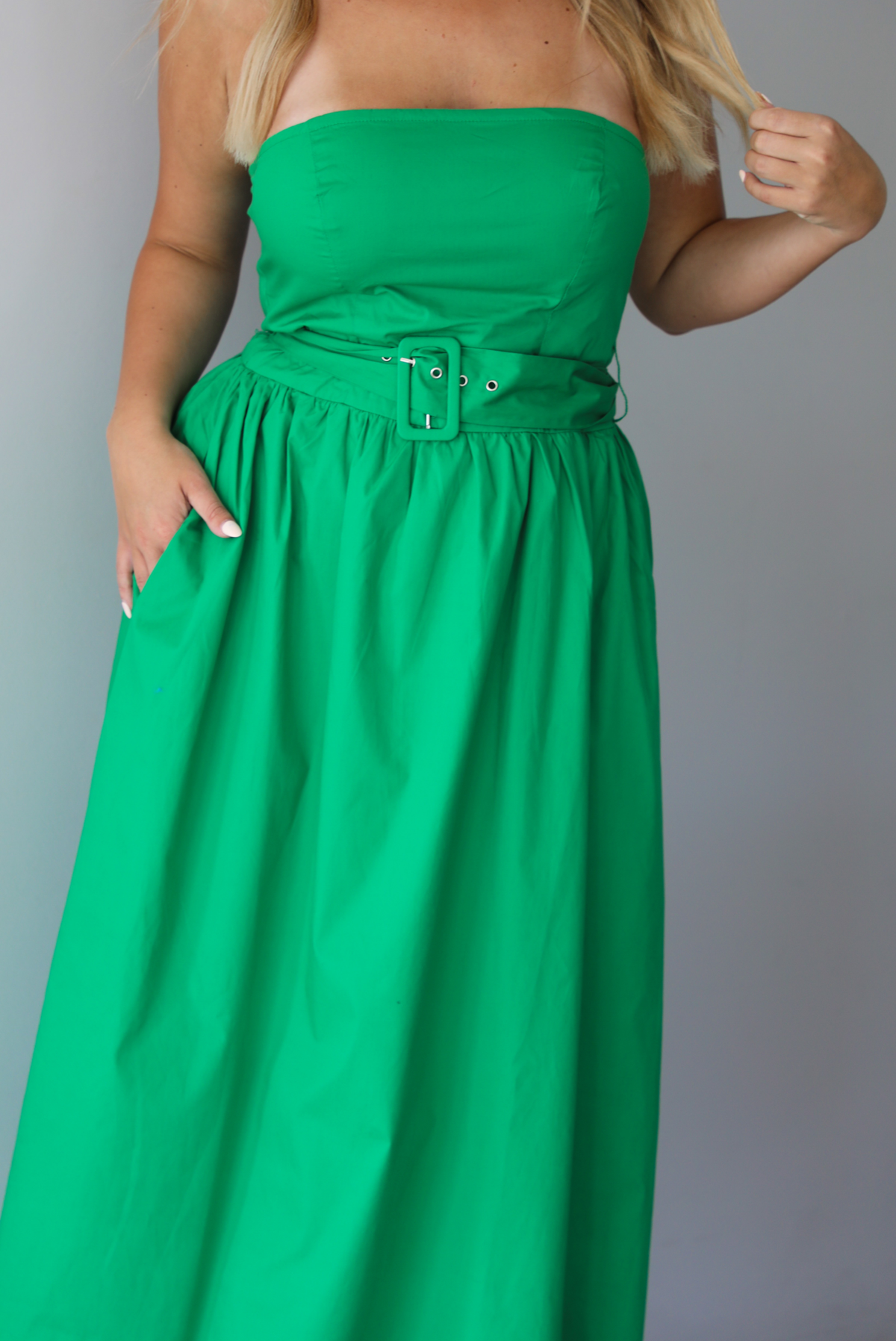 Woman wearing a green strapless midi dress that has a fitted bodice, belted waistline detail, functional pockets on either side, and a midi length indoors against a white wall.