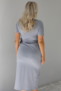 Woman wearing a grey midi length t-shirt textured dress that has short sleeves that cuff slightly at the hem, a tie detail just under the bodice on the left side, and a split hem detail on the left side. The photo shows a closeup of the back of the dress. The photo is taken indoors against a white wall with white marble flooring. 