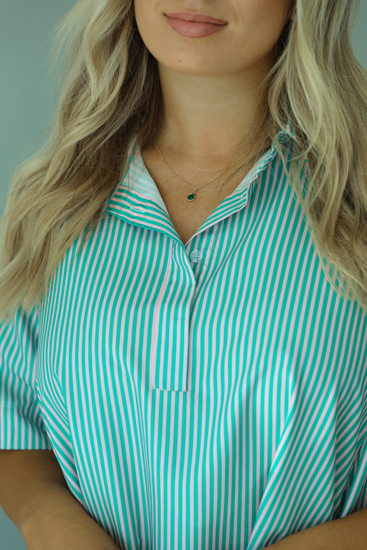 Woman wearing a green and pink striped dress that has a collared neckline, functional quarter button closures in the front, and a flowy skirt. The photo is taken indoors against a white wall and shows a closeup of the collared neckline and the few button closures at the top of the bodice. 