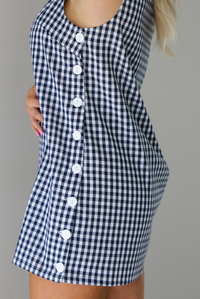 Woman wearing a navy and white gingham print patterned dress that has non-functional button closures on the left side. The photo is taken indoors against a white wall. The photo shows the button closures on the side. 
