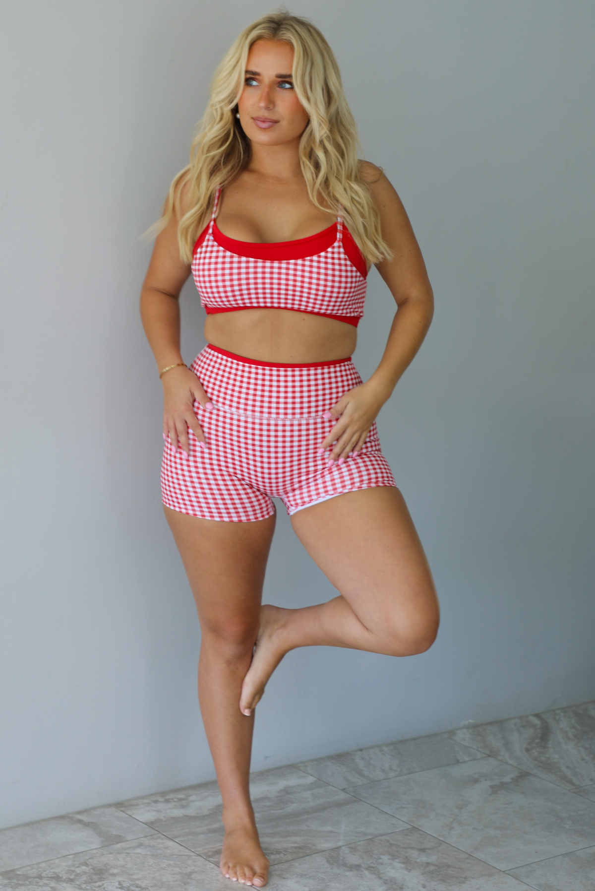 A woman modeling a red and white gingham two-piece athletic set featuring a sports bra and high waisted biker shorts with solid red trim indoors against a white wall with white marble flooring. 