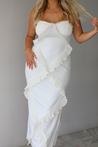 Woman wearing a white maxi length dress with ruffled details along the front indoors against a white wall with white marble flooring. 