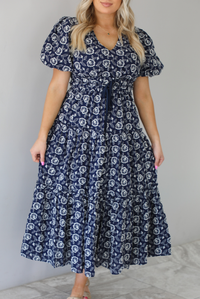 Woman wearing a navy dress that has a white embroidered floral print pattern throughout, a cinched waistline, and flowy maxi length skirt. The photo shows a closeup of the dress. The photo is taken indoors against a white wall.