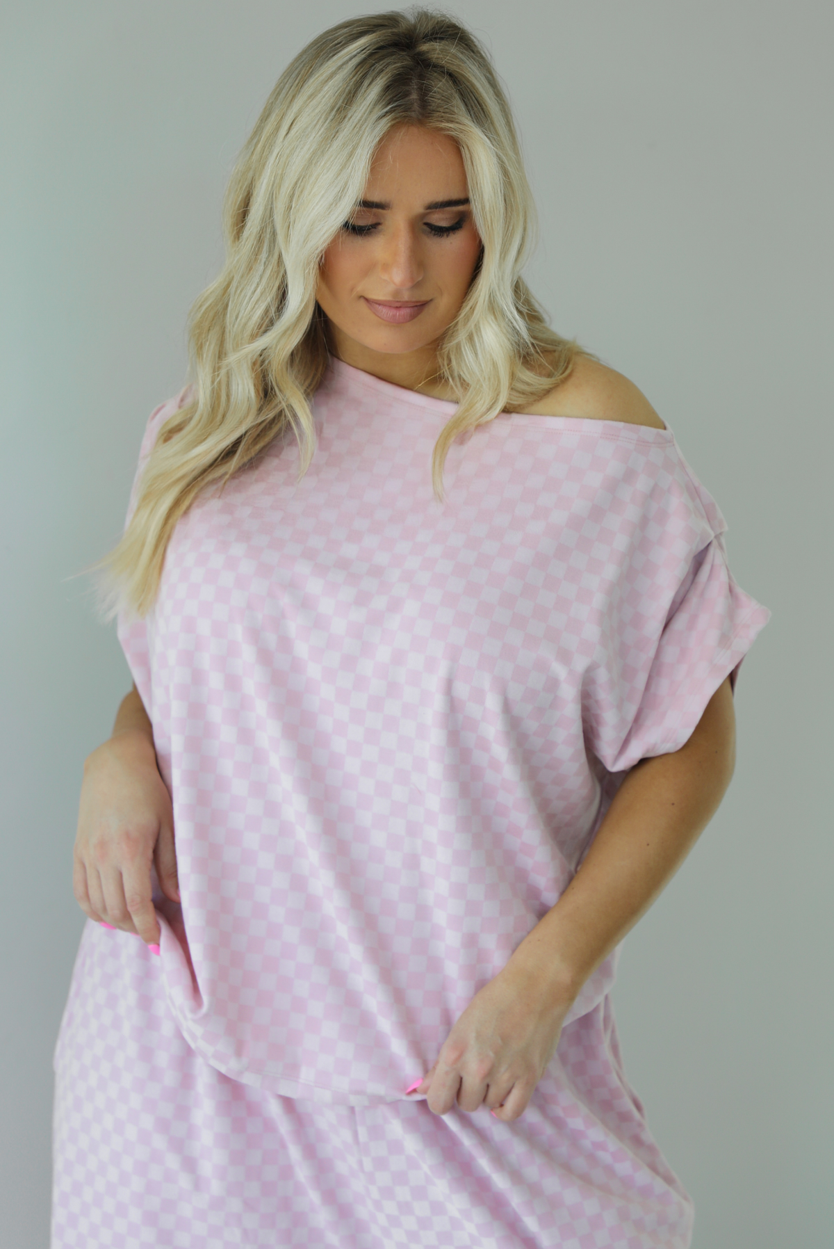 Woman wearing a pink checkered set featuring a slouchy top and pants. The photo is taken indoors against a white wall and shows a closeup of the top, which is worn off the shoulders and has a relaxed fit, and is untucked to show the full length. 