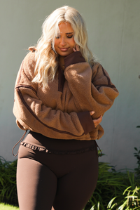 Woman model wearing a tan and brown quarter zip sweater with dark brown athleisure pants. The photo is taken outdoors against some greenery and a white wall. The photo showcases the sweater close up. 