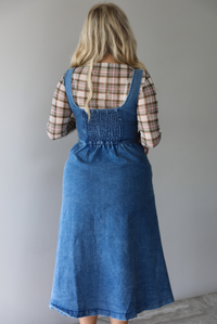 A woman shown from behind wearing a medium wash denim midi dress with button closures along the  front, with pockets on either side, paired with a sheer, multi-colored plaid long sleeved top underneath, standing indoors against a white wall with white marble flooring. The back shows a smocked detail. 